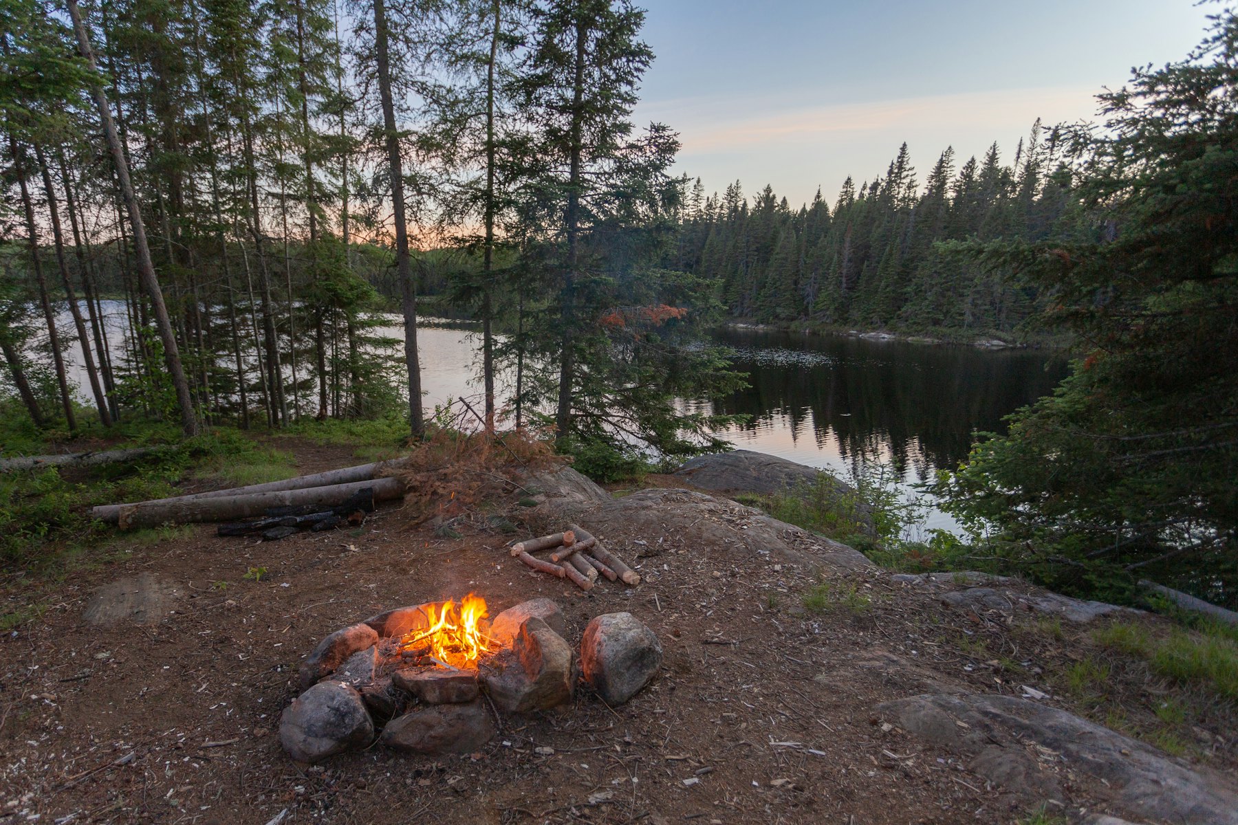 A small fire in a stone ring on a granite point at dusk, tall pines behind, a still lake beyond.