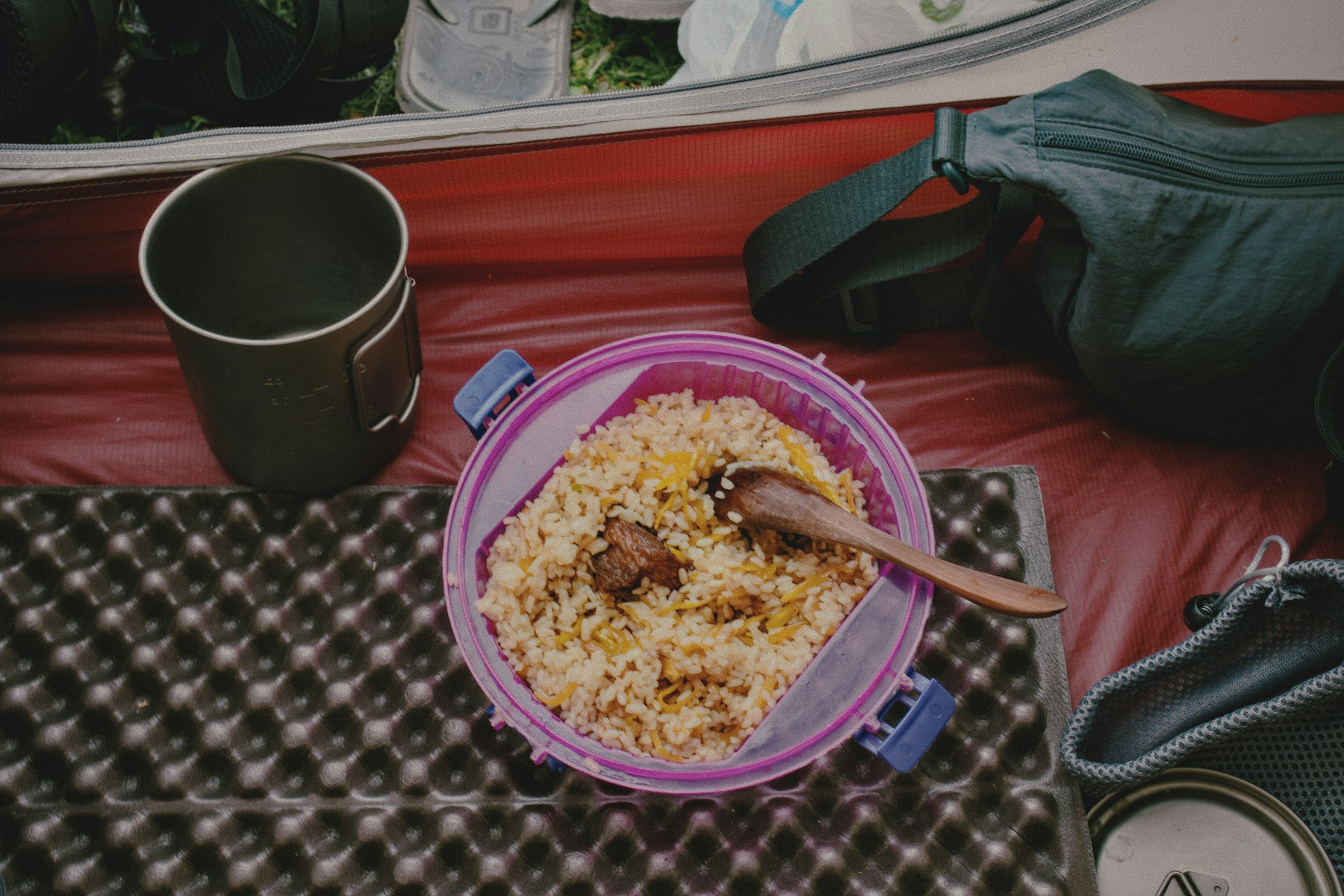 A pot of rice and meat with a wooden spoon in it, on a foam pad inside a tent, a steel mug beside it.