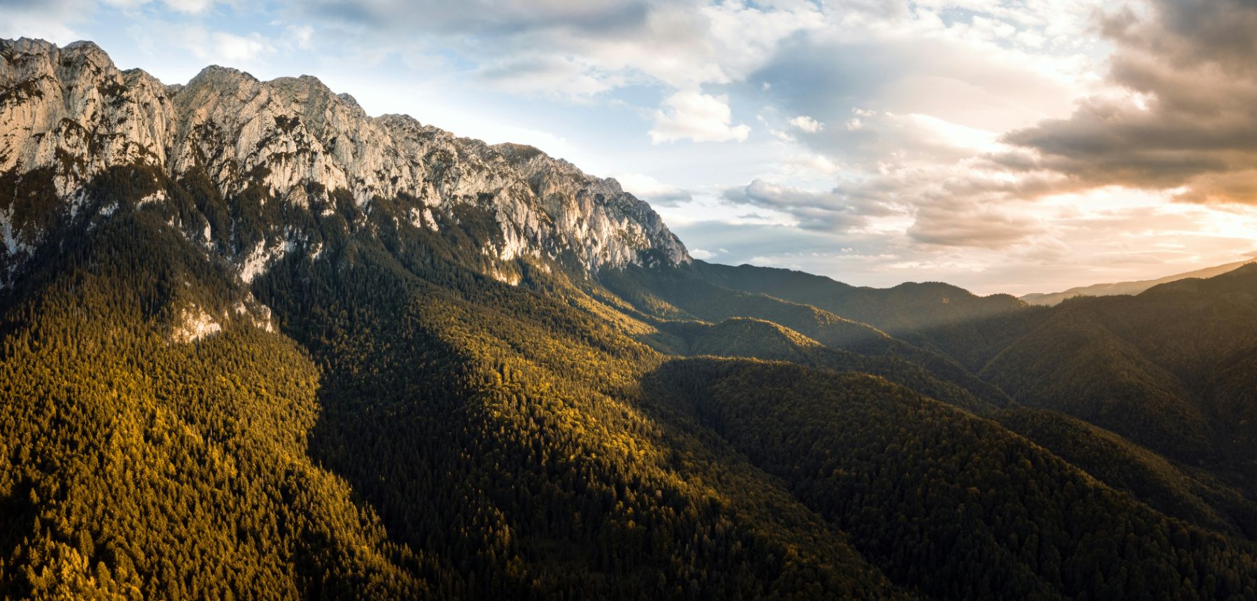 A long limestone ridge above dark forested slopes, the evening sun catching the treetops and the far valleys fading into haze.