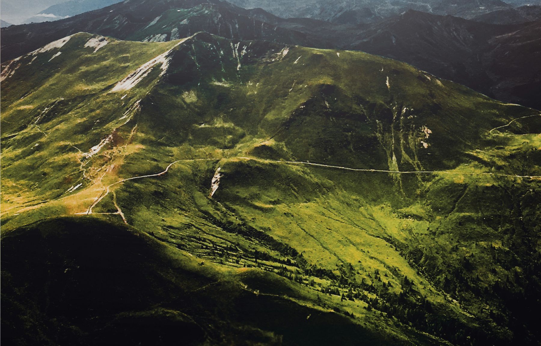 A trail winding along the crest of a green alpine ridge, sunlit meadows falling away on both sides and ridge after ridge fading into haze behind it.