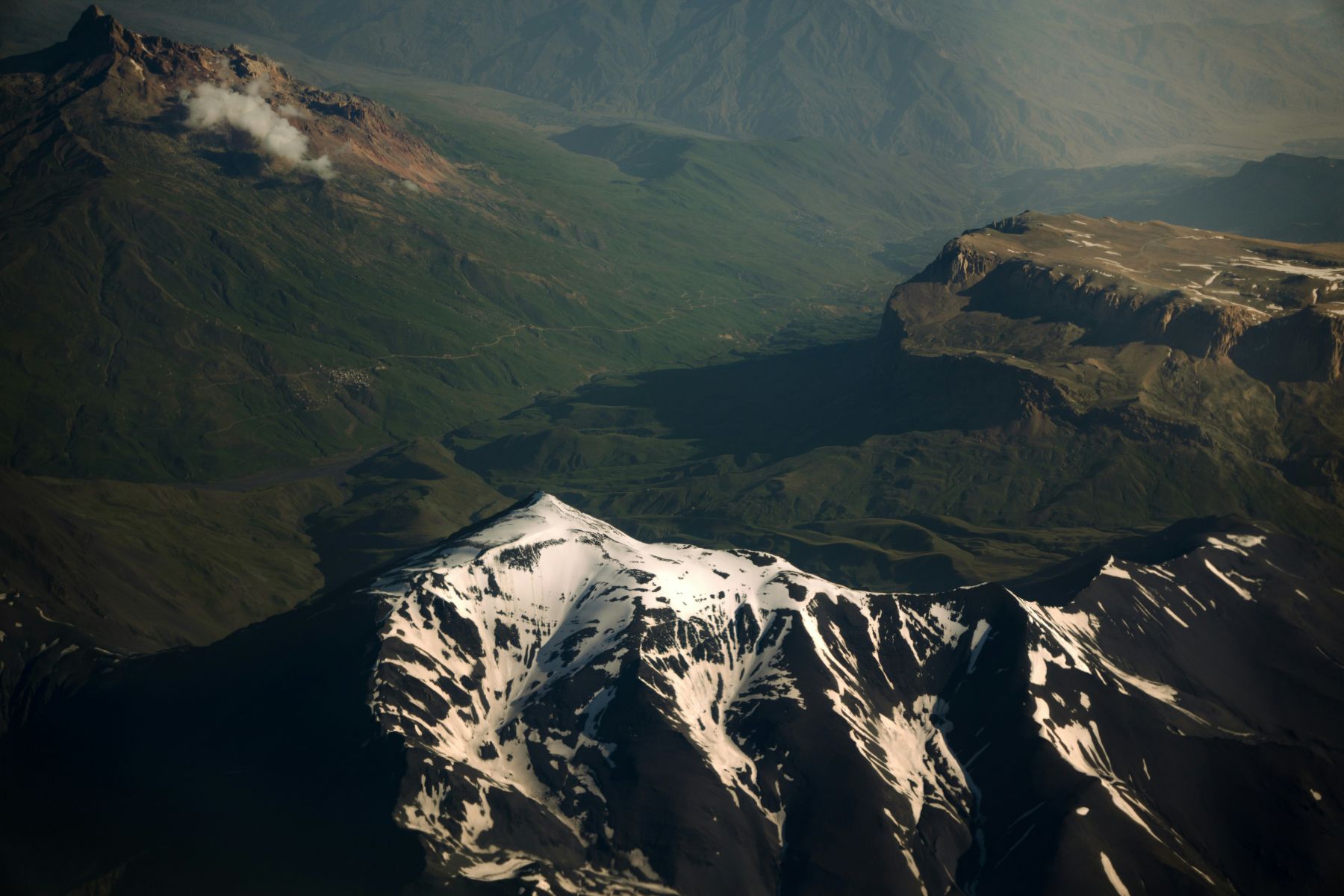 From the air: a snow-streaked peak in the foreground and, behind it, green valleys with a road threading between them toward a distant plateau.