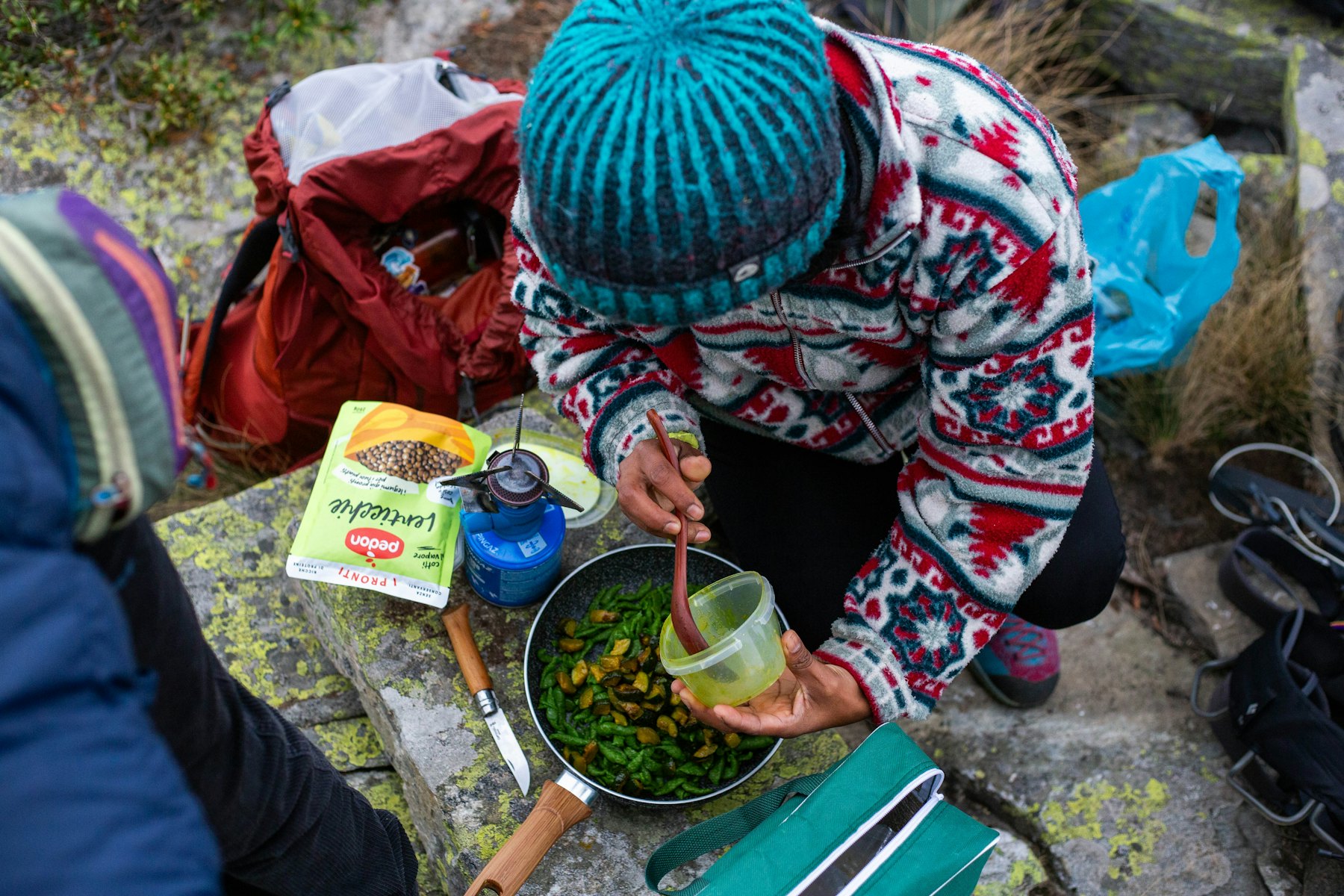 A hiker in a patterned sweater crouched on lichen-covered granite, stirring a pot on a small canister stove beside their pack.