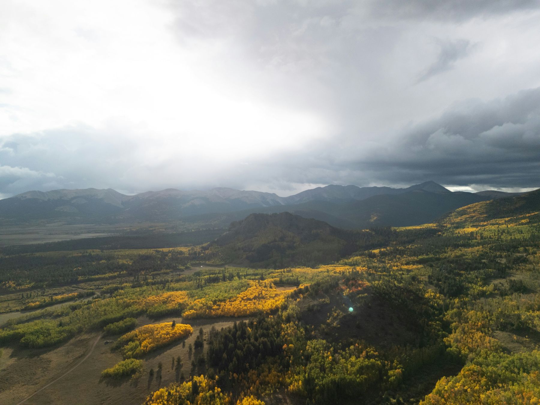 A broad valley of aspen in yellow under a heavy storm sky, a track running out across the flats toward a dark ridge and the mountains beyond.